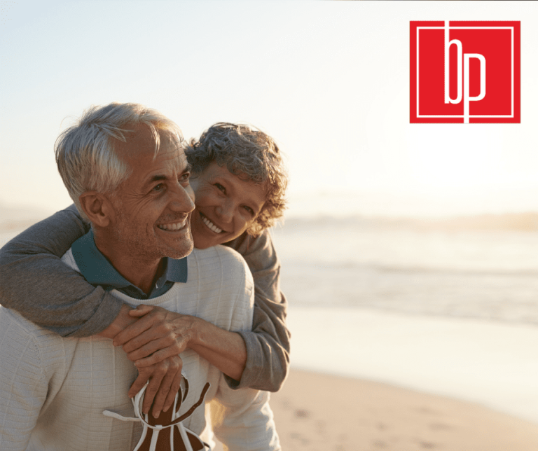 An older woman with her arms wrapped around her husband as they walk along the beach